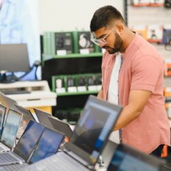 A young man stands behind his laptop at the electronics store.